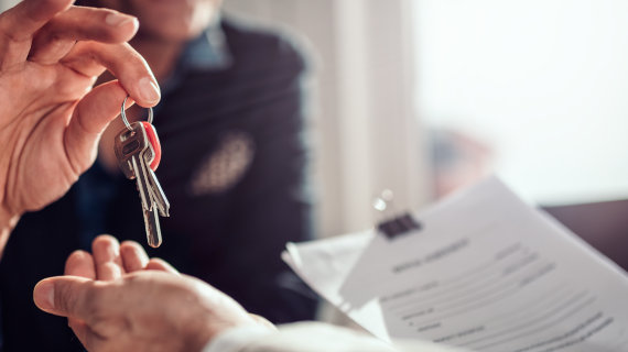 Nahaufnahme von zwei Händen beim Übergeben eines Schlüsselbunds. Eine Hand hält die Schlüssel am Schlüsselring, die andere greift danach. Im Hintergrund ist ein teilweise sichtbares Dokument auf einem Klemmbrett zu sehen.